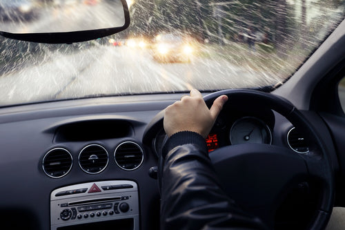View from inside a car driving in rainy weather with water splashing on the windshield and headlights of oncoming vehicles visible.