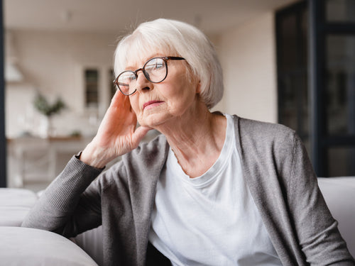 Elderly person wearing glasses and a gray cardigan, sitting on a couch in a softly lit room, resting their head on their hand.