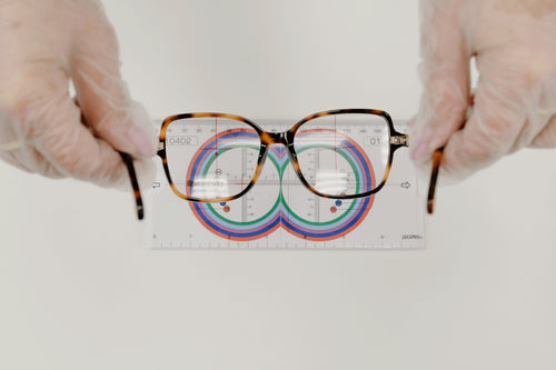 Hands wearing gloves holding eyeglasses over a lens meter chart for optical measurement and lens fitting.