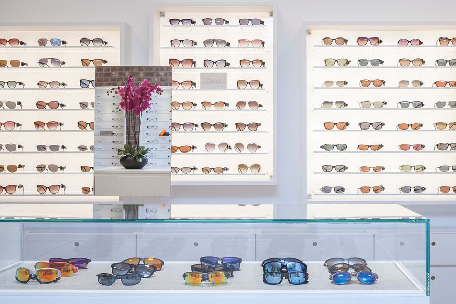 Display of various sunglasses and eyeglasses on illuminated wall shelves and glass countertop in a modern eyewear store.