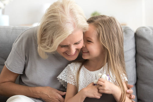 Child holding a small toy and gift box, sharing a joyful moment with an older woman on a grey couch in a bright living room.