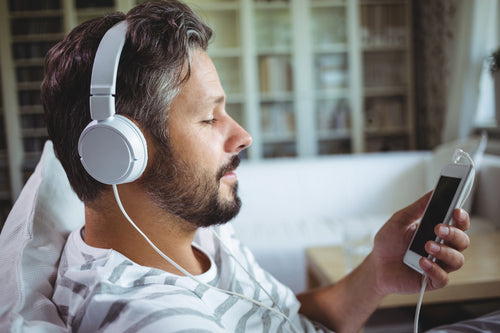 Man relaxing on a couch listening to music with white headphones connected to a smartphone in a cozy living room.