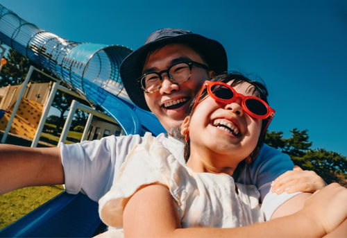 A father and his daughter laughing together, with a playground slide in the background.