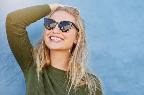 Smiling person wearing round sunglasses and hoop earrings, posing in front of a light blue textured wall with one hand on their head.